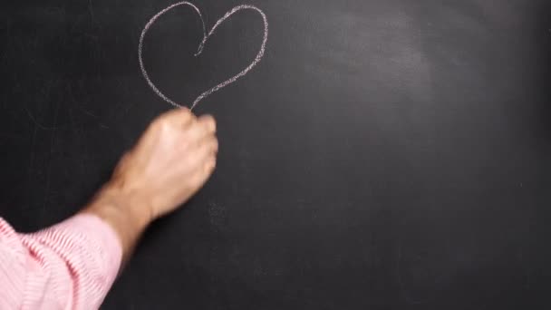 A man in a shirt draws two men and a heart with chalk on the blackboard. Valentines Day holiday concept. — Stock Video