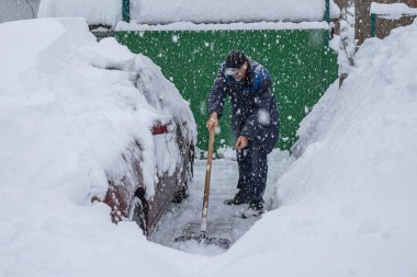 Bir adam evinin önündeki sokaktan kar kürüyor. Yakınlarda karla kaplı bir araba var. Arka plandaki yeşil kapıda. Büyük kar taneleri düşüyor. Kar fırtınası sırasında sokakları temizlemek