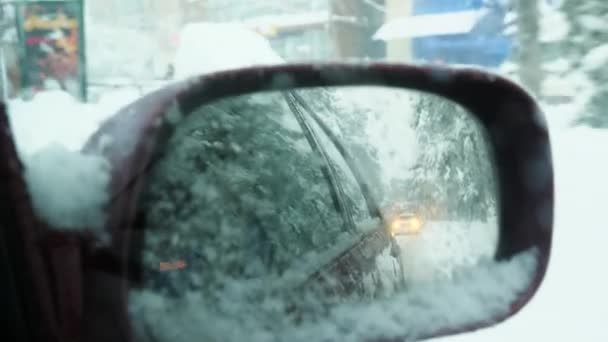 Widok z bliska z wnętrza w lusterku wstecznym bocznym samochodu. Samochód przejeżdża przez śnieżne miasto. Światła samochodów, drzewa i opady śniegu odbijają się w lustrze.. — Wideo stockowe