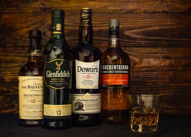 4 şişe premium 12 yıllık viski, Balvenie, Auchentoshan, Glenfiddich, Dewars ve koyu ahşap arka planda bir bardak buz. Rusya, Krasnodar, 11 Aralık 2021