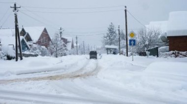 Araba karla kaplı bir yolda gidiyor. Arkadan bak. Yol kenarlarında sürüklenme izleri var. Evet. İnsanlar otobüs durağında duruyor. Arka planda, gri kış gökyüzü ve kar yağışı