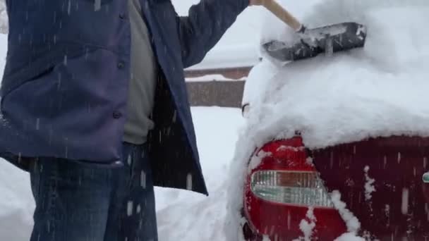 一个穿着夹克和牛仔裤的男人正在把雪从他的车上铲走 暴风雪中一辆红色的汽车被雪覆盖着 背景中的降雪 — 图库视频影像