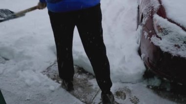 Mavi ceketli ve siyah pantolonlu bir adam evinin önünde kar kürüyor. Yakınlarda kırmızı bir araba duruyor. Gökten kar yağıyor.