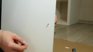 Beyaz bir dolap kapısının kulpunu beceren bir adamın yakın çekimi. Arka planda karton ve kaplama var. Evde mobilyaları kendi ellerinle monte ediyorsun.