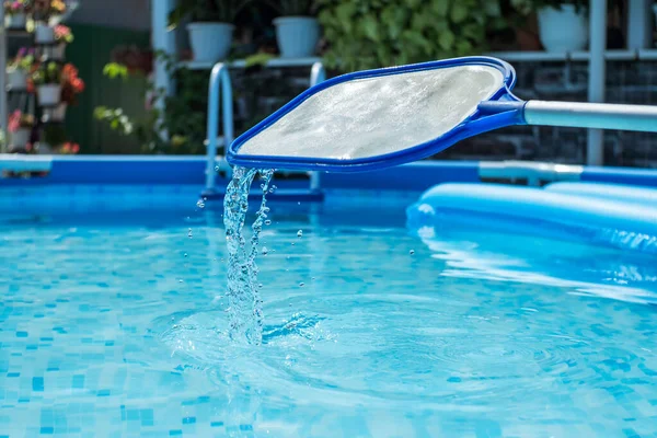 清澈蓝水的背景下 用水滴清洁游泳池网 背景是一个种满鲜花的庭院 清洁树叶和昆虫的圆形框架池和网状滑板 — 图库照片