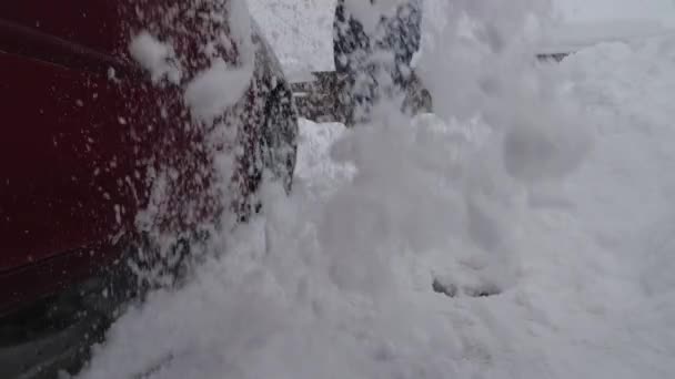 ジャケットとジーンズの男が赤い車の隣に雪を掘っている 接近中だ 背景には 雪の厚い層で覆われた家の茶色の柵と屋根があります 空から雪が降る — ストック動画