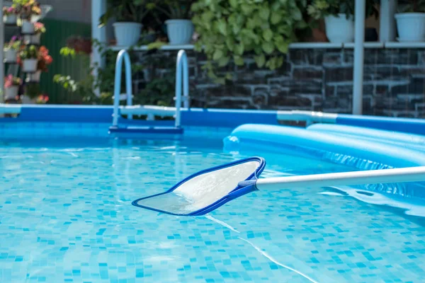 Net-skimmer in a clear water pool with an inflatable mattress. Backyard with flowers in the background. Round frame pool and leaf netting skimmer for cleaning leaves from the surface of the water