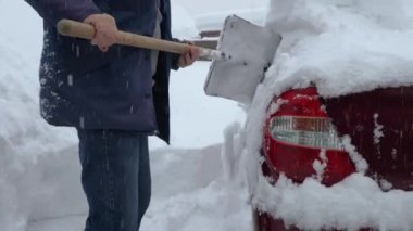 Ceketli ve kotlu bir adam kırmızı bir arabadan kar kürüyor. Kar fırtınası sırasında araba kar altında kaldı. Gökten kar yağıyor.