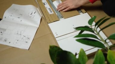 Bir adamın elleri raflı bir kutuyu açıyor. Ön tutsağın üzerinde bir çiçek var. Yerde talimatlar, kartonlar ve aletler var. Evde kendi elleriyle mobilya montajı yapıyor.