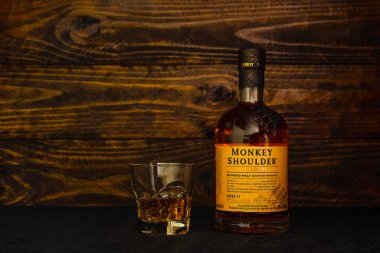 Bir şişe Monkey Shoulder Scotch Whiskey ve koyu ahşap arka planda buzlu bir bardak. Viski, üç damıtıcıdan karıştırılmış malt ile yaratıldı. Rusya, Krasnodar, 11 Aralık 2021