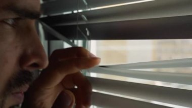 Perdelerdeki yarıktan dışarı bakan sakallı bir adamın yakın çekimi. Gizli izleme