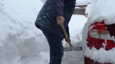 Şapkalı, ceketli ve kot pantolonlu bir adam evinin yakınındaki bölgeden kar kürüyor. Yakınlarda kar fırtınası sırasında karla kaplı kırmızı bir araba var. Arkaplanda kar yağışı