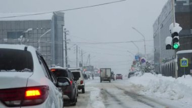 Arabalar trafik ışığında karla kaplı bir kavşakta duruyor. Yeşil ışık yanıp sönüyor ve sarı ışık yanıyor. Evin arka planında, gri bir kış gökyüzü ve kar yağışı.