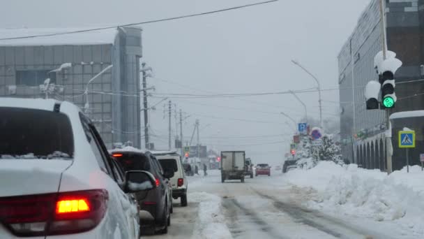 Samochody stoją na pokrytym śniegiem skrzyżowaniu na światłach. Zielone światło miga i zapala się żółte światło. W tle domu, szare zimowe niebo i opady śniegu. — Wideo stockowe