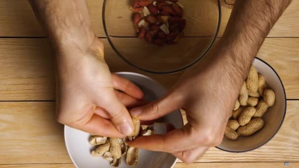 Vista dall'alto mani maschili sbucciare arachidi arrosto dalle loro conchiglie. Primo piano. Sfondo in legno. Sbucciare un'arachide terrosa a casa. — Video Stock