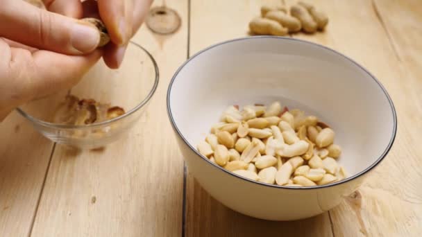 Des images au ralenti d'un homme épluchant des cacahuètes de leurs coquilles, jetant des noix et des coquilles dans différentes tasses. Une poignée d'arachides reposent sur une table en bois. Peler une cacahuète terreuse à la maison. — Video