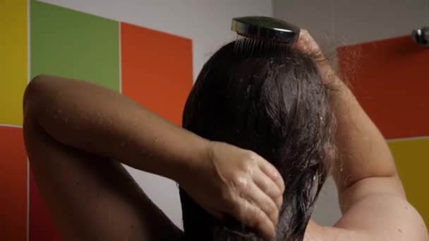 Közelkép egy nőről, aki a hosszú, sötét haját vizeli egy zuhanyozó géppel. Színes csempék vannak a háttérben. Visszanézni. Hajápolási koncepció