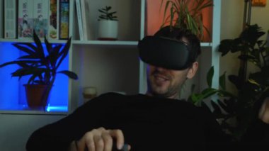 Siyah kazaklı traşsız bir adam karanlık bir odada otururken sanal gerçeklik gözlükleriyle dans ediyor. VR kulaklık kullanıcısı, geleceğin eğlencesi. Krasnodar, 16 Şubat 2022