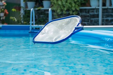 Mavi berrak suyun arka planında su damlacıkları olan havuz temizleme ağı. Arka planda çiçekli bir avlu var. Yuvarlak çerçeve havuzu ve yaprakları ve böcekleri temizlemek için ağ süzgeci