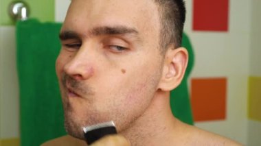 Beyaz bir adam evinde, banyoda elektrikli tıraş makinesiyle tıraş oluyor. Bir adam sakalını tıraş ediyor ve yüzünü kareliyor. Arka planda çok renkli fayanslar var. Erkek güzelliği kavramı