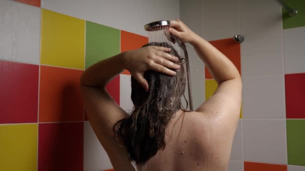 一个女人正在用淋浴间喷出的水来冲洗她长长的黑发上的洗发水 后视镜后面的瓷砖色彩斑斓 头发护理和个人卫生的概念 — 图库视频影像