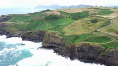 Dalgalı bir denizin üzerindeki yeşil doğal alandaki kayalık bir sahil uçurumunun havadan görünüşü. Ada, Cantabria, İspanya. 4K