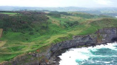 Dalgalı bir denizin üzerindeki yeşil doğal alandaki kayalık bir sahil uçurumunun havadan görünüşü. Ada, Cantabria, İspanya. 4K