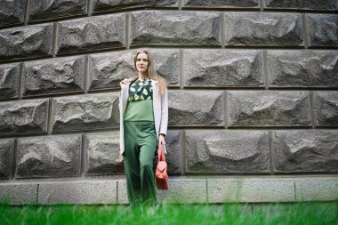 Genç güzel kız büyük tuğlalardan oluşan büyük bir duvarın yanında duruyor. Güzel bir kadın öğrenci olmak ya da bir iş bulmak ister. Yeşil günlük kıyafet, turuncu çanta ve aksesuarlar. Yaşam tarzı kentsel, şehir yürüyüşü