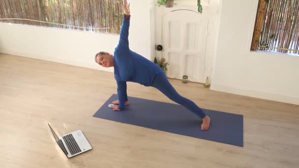 这位身穿蓝色衣服的瑜伽老师一边做伸展运动 一边看她的笔记本电脑 一边在家里给她的学生们上网络课 — 图库视频影像