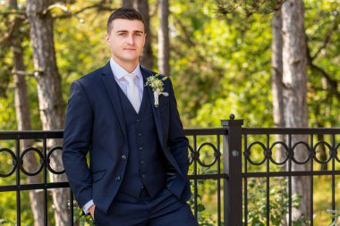 Zarif lacivert takım elbiseli yakışıklı damat açık hava fotoğraflarına bakıyor. Şık damat modası.