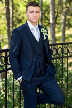 Zarif lacivert takım elbiseli yakışıklı damat açık hava fotoğraflarına bakıyor. Şık damat modası.