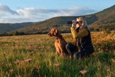 Genç bir dişi avcı, güneşli bir sonbahar akşamında yanında Macar Vizsla köpeği ile kuş gözlemlemek için dürbün kullanıyor. Bir av köpeğiyle avlanmak..