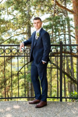 Zarif lacivert takım elbiseli yakışıklı damat açık hava fotoğraflarına bakıyor. Şık damat modası.