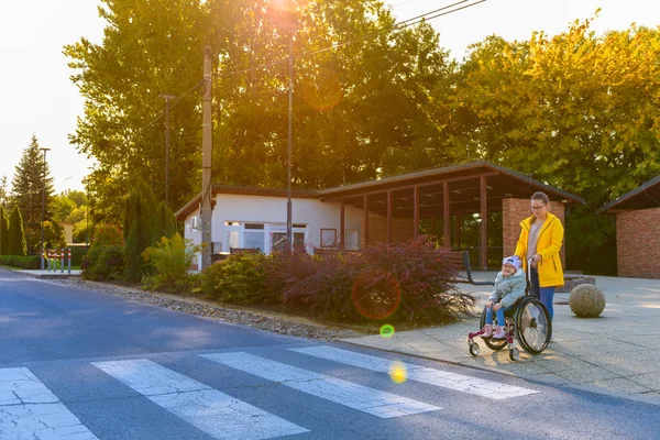 脳性麻痺の少女と一緒に車椅子を押している若い母親は 治療への道を渡ります — ストック写真