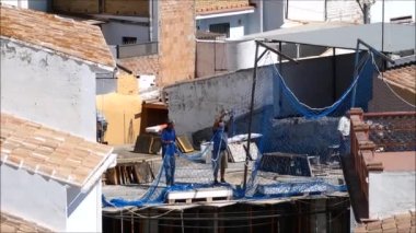 Alora, İspanya - 14 Temmuz 2019: Şehrin iç kesimlerinde mavi güvenlik ağı kurmaya çalışan iki işçi. Yeni inşaat alanındaki işçiler, kahverengi köyün üzerine mavi ağ örtüsü dikiyorlar..