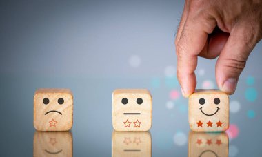 A man uses his finger to select a facial grin emoticon to place on a wood block. A user experience or customer satisfaction survey, poll, or questionnaire. The customer is the concept.