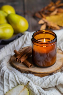 Koyu cam bir kavanozda kokulu bir mum ve tarçın çubukları bir süveterin üzerinde duruyor, arka planda yeşil armutlar.