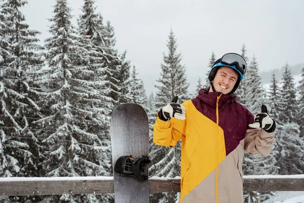 Homem em equipamento de esqui, usando óculos de segurança mostra polegares para cima, fica contra a montanha e árvores. Esportes de inverno e recreação, lazer atividades ao ar livre — Fotografia de Stock