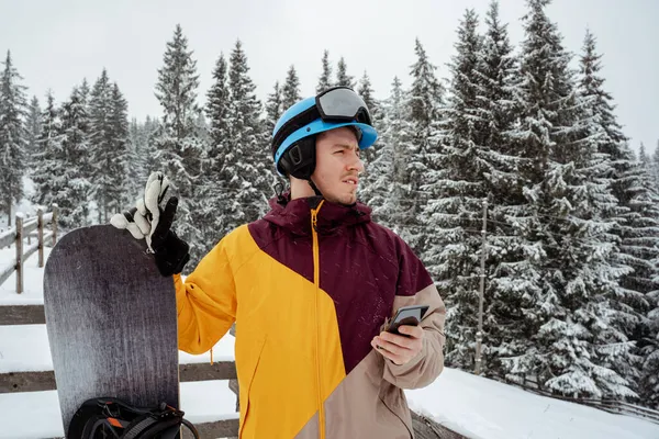 Winter sport and recreation, leisure outdoor activities. Man in ski equipment, wearing safety glasses with smartphone . Snowboarder man hiking at mountain