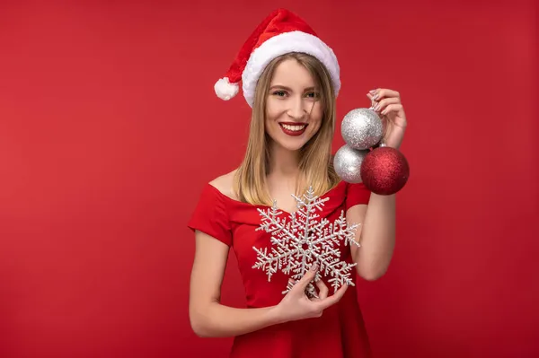 Фотографія привабливої жінки в різдвяному капелюсі та червоній сукні, містить різдвяні іграшки та щасливу посмішку. Ізольовані на червоному тлі — стокове фото