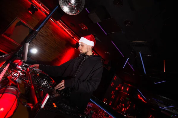 Nowy Rok w nocnym klubie. Wesołych Świąt Młody, pewny siebie DJ w czerwonym kapeluszu Świętego Mikołaja mieszając muzykę na gramofonach na przyjęciu świątecznym. Świecąca kula disco. Czarno-czerwone tło klubu. — Zdjęcie stockowe