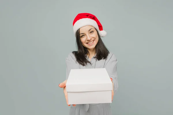 Foto de linda hembra sonriente en sombrero de Navidad dando un regalo. Lleva camisa gris, fondo gris aislado Fotos De Stock