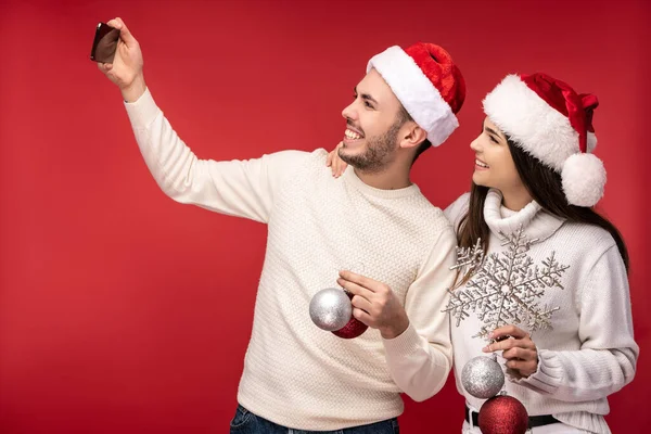 Foto de pareja dulce en sombreros de Navidad y con juguetes de Navidad, se ve feliz. Hombre y mujer se toman una selfie y sonríen, aislados sobre fondo rojo Imagen De Stock