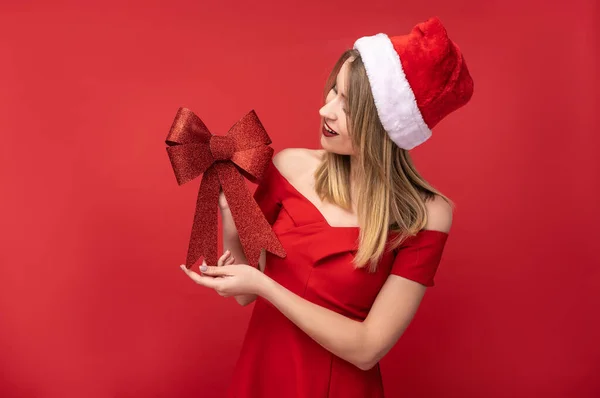 Foto de mujer atractiva en sombrero de Navidad y vestido rojo, sostiene el lazo rojo para el árbol de Navidad y míralo. Aislado sobre fondo rojo Imagen De Stock