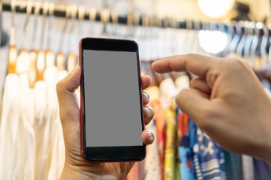 Kadın giyim mağazasında ve elinde telefon, ahşap askılarda giysiler, online alışveriş konsepti.