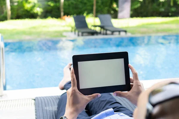 Business man lying on a chair at the swimming pool, holding a tablet with blank screen.