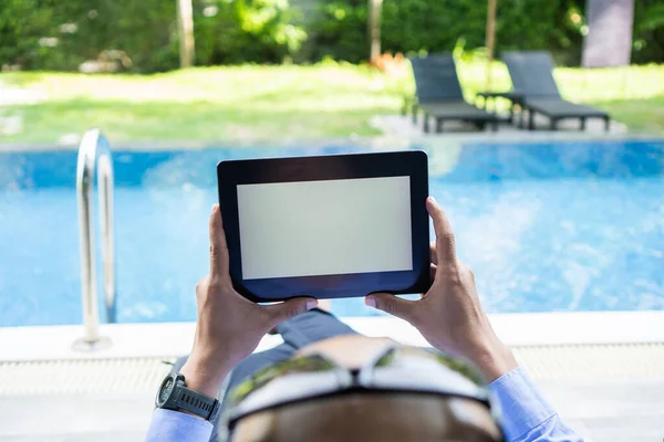 Business man holds tablet with isolated screen in his hands, blank white screen at swimming pool.