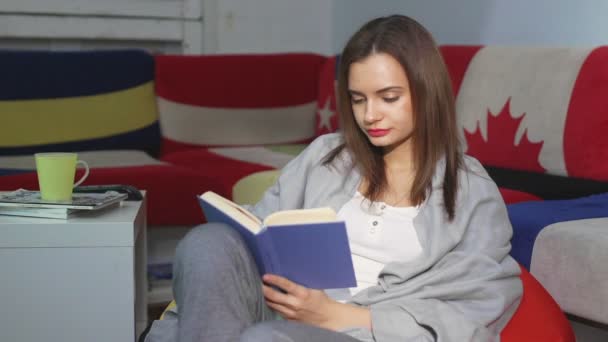 A young woman is sitting at home on an ottoman and reading a book. A woman is resting indoors on a comfortable ottoman in her free time. Have a nice free time on the weekend — Stock Video