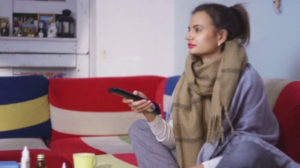 Боковой вид: больная молодая женщина сидит на диване перед различными таблетками на столе, смотрит телевизор — стоковое видео
