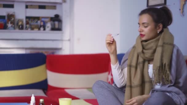 Side view a young woman feels unwell and takes her temperature. A young woman sitting on the sofa, with a scarf, takes her temperature. Drinks the medicine — Stock Video